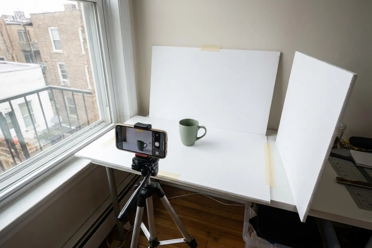 DIY product photography setup: white foam boards around a small table beside a window, with a phone on a tripod aimed at a sage green mug